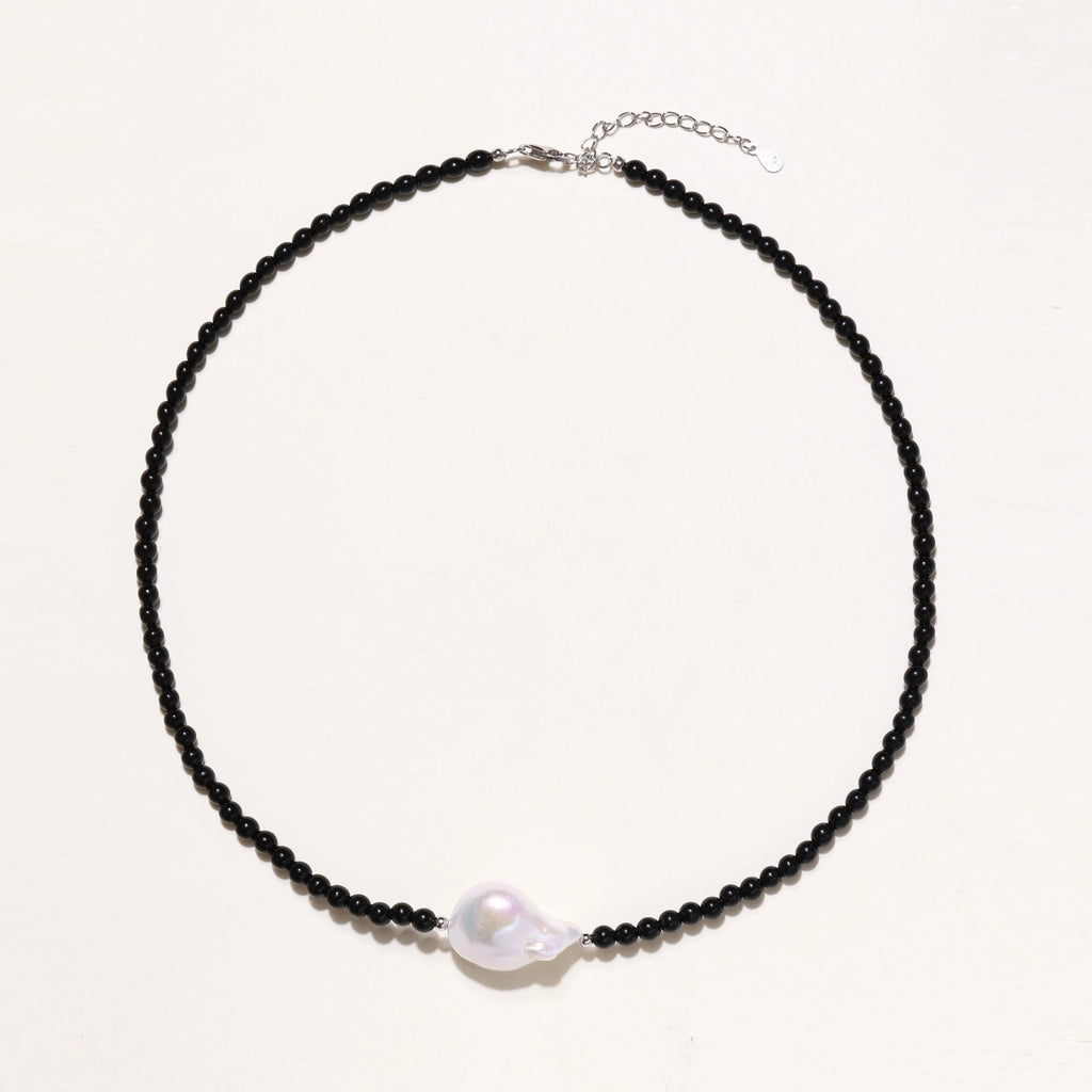 Beaded black agate necklace with iridescent baroque freshwater pearl centerpiece on cream background.