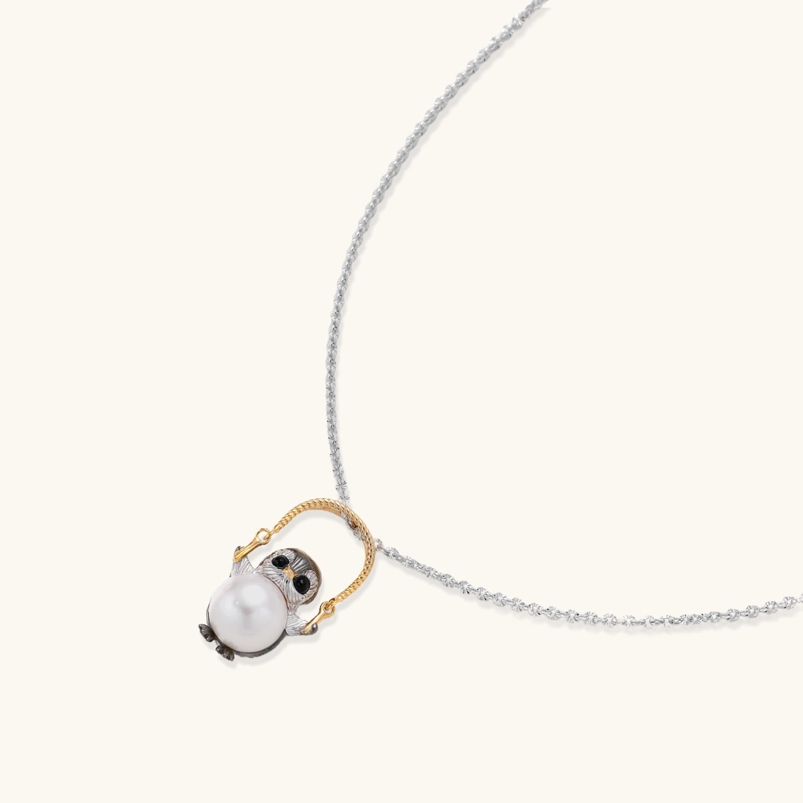 Whimsical penguin pendant holding a white pearl on a gold swing, rhodium-plated silver chain on cream background.