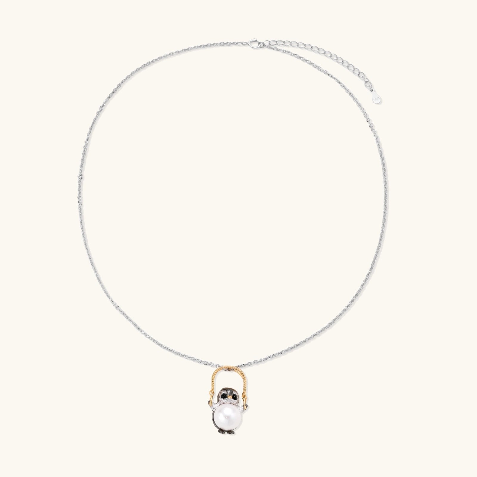 Whimsical penguin pendant holding a white pearl on a gold swing, rhodium-plated silver chain on cream background.
